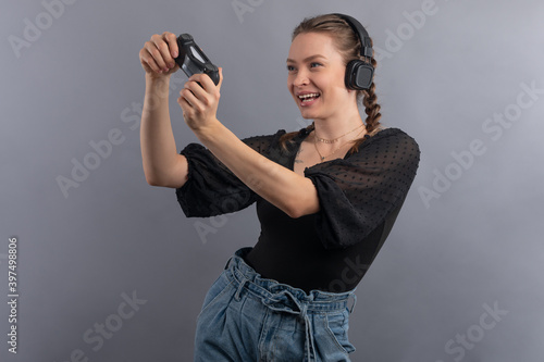 Close-up view of emotional smiling gamer woman playing video game and using joystick and headphones on grey background. Gambler concept. Using modern technology.