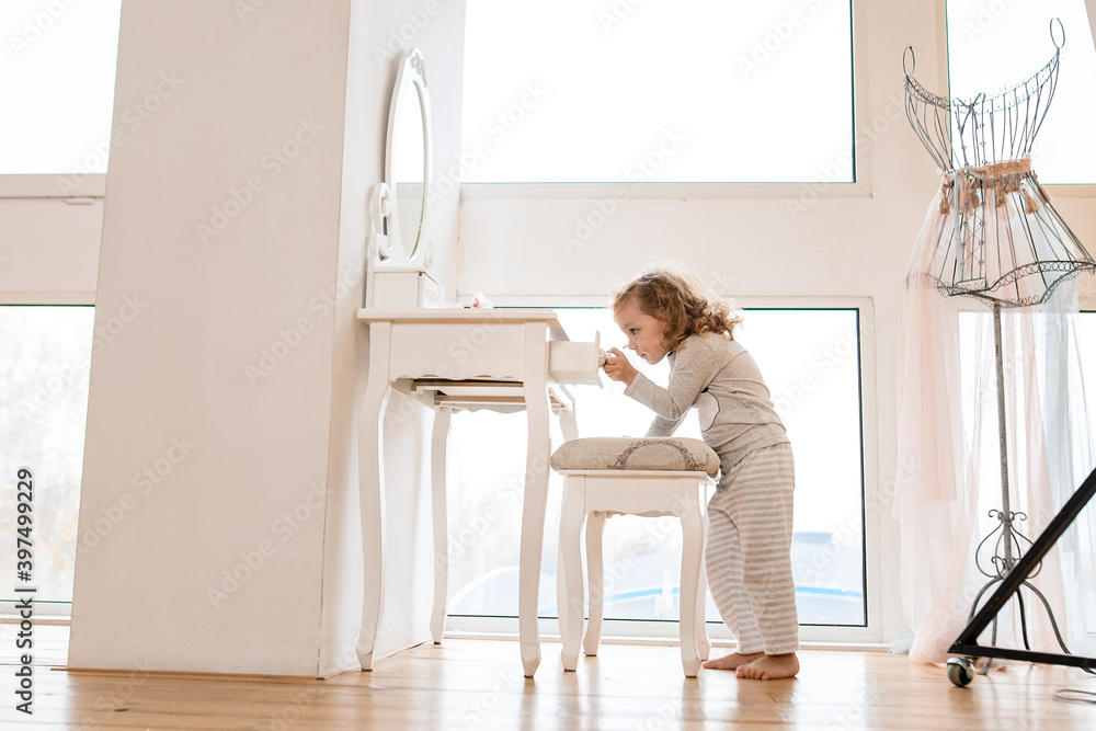 Handsome little girl stands near console table with mirror and pulls drawer out of it Handsome little girl stands near console table with mirror and pulls drawer out of it
