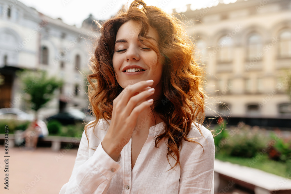 Smiling lovable charming lady with curly red hair smiling with closed eyes in sunlight on background of old street