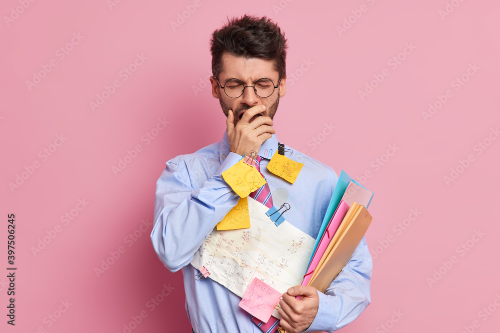 Tired student has sleepy expression covers mouth with hand and yawns dressed in formal clothes prepares for exams poses against pink background. Executive worker makes memo notes for project Tired student has sleepy expression covers mouth with hand and yawns dressed in formal clothes prepares for exams poses against pink background. Executive worker makes memo notes for project