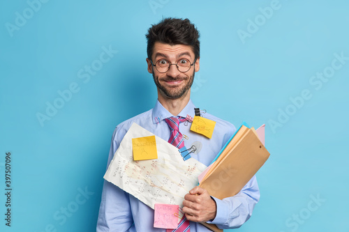 Cheerful bearded male financier prepares stratup project poses with papers and folders dressed in formal clothes wears round spectacles isolated over blue background. Office worker being busy photo