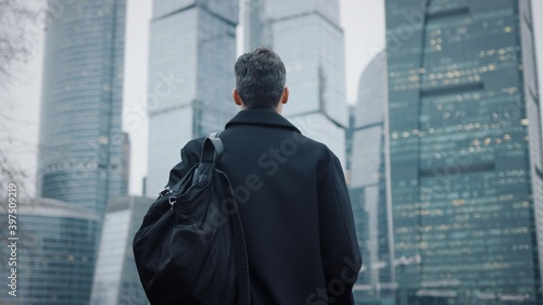 Man in coat looking at the skyscrapers pan shot right to left. Gimbal back shot of businessman in eyeglasses near skyscrapers in business city