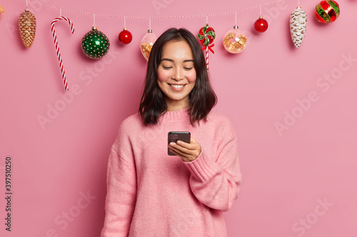 Obraz na plátně Photo of delighted brunette Asian woman with eastern appearance holds modern mobile phone sends messages of congratulation on New Years Eve wears casual jumper poses against christmas decor