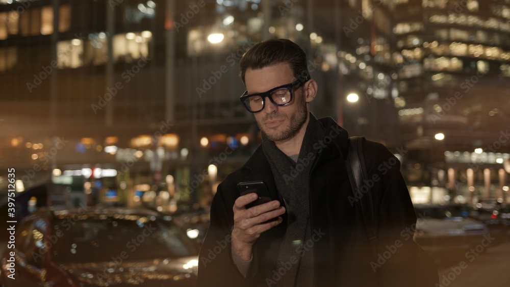 Man in eyeglasses with phone in hand on background of building with lights on. Gimbal night shot of caucasian man texting in phone near business building Man in eyeglasses with phone in hand on background of building with lights on. Gimbal night shot of caucasian man texting in phone near business building