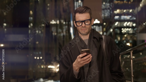 Man in eyeglasses with phone in hand on background of building with lights on. Handheld night shot of caucasian man texting in phone near business building with cars