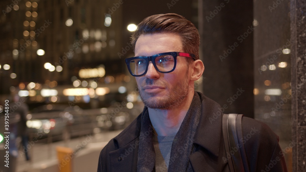 Man in eyeglasses standing on background of business skyscrapers, windows with lights. Portrait gimbal shot of businessman in coat standing near business centre downtown at night Man in eyeglasses standing on background of business skyscrapers, windows with lights. Portrait gimbal shot of businessman in coat standing near business centre downtown at night
