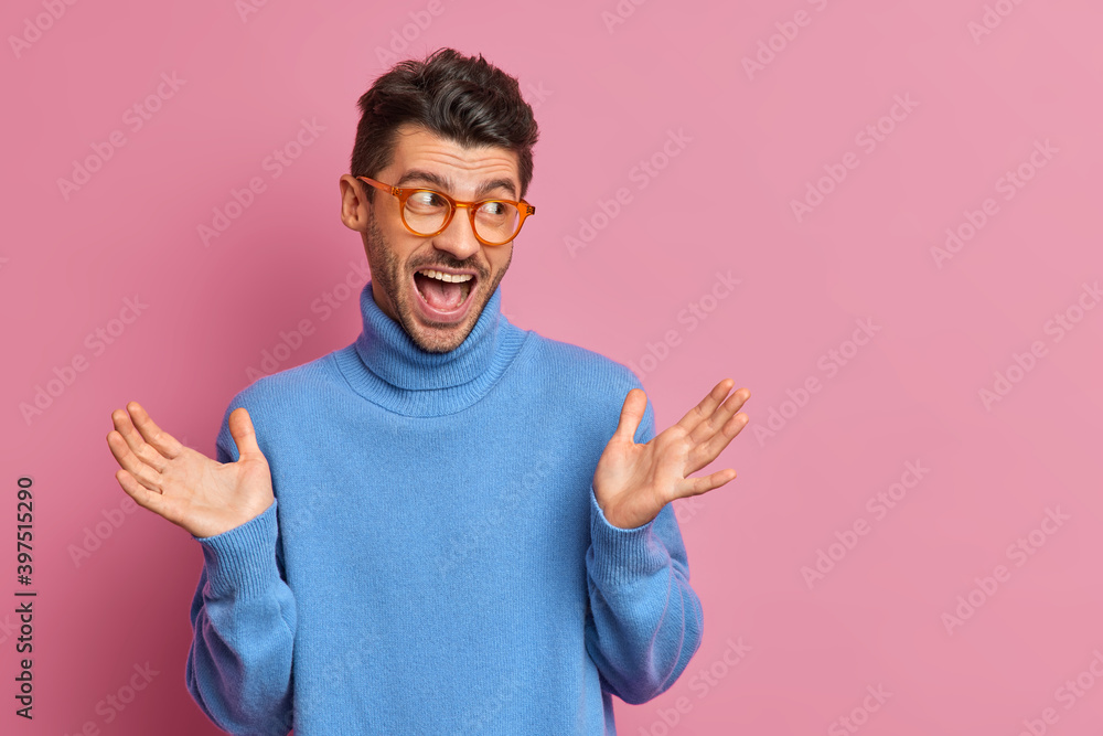 Optimistic hipster guy raises palms and reacts on awesome news dressed in casual blue poloneck looks gladfully aside poses against pink background with copy space for your promotional content Optimistic hipster guy raises palms and reacts on awesome news dressed in casual blue poloneck looks gladfully aside poses against pink background with copy space for your promotional content