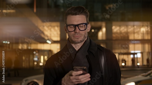 Man in eyeglasses with phone in hand on background of building with lights on. Gimbal night shot of caucasian man texting in phone near business building