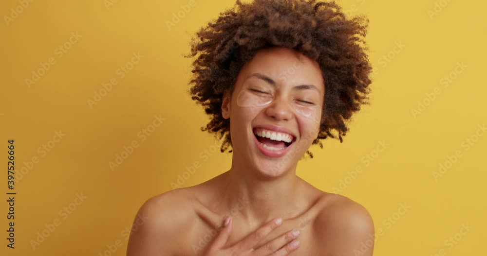 Pretty mixed race young woman has overjoyed expression wears hydrogel under eye patches for beautiful face skin laughs joyfully poses naked against yellow background. Beauty treatment concept