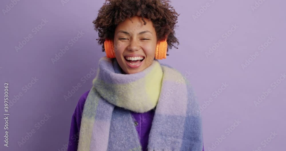 Overjoyed curly haired woman expresses happiness has funny talk and laughs positively wears warm winter scarf around neck listens favorite music via headphones isolated over purple background Overjoyed curly haired woman expresses happiness has funny talk and laughs positively wears warm winter scarf around neck listens favorite music via headphones isolated over purple background