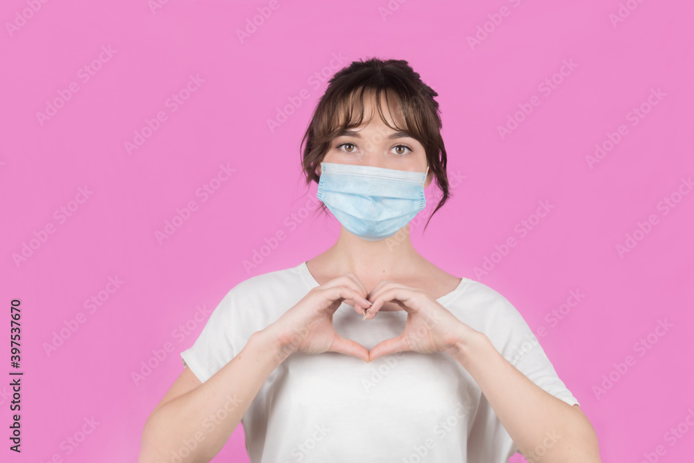 Portrait of a young woman in a medical mask making a heart gesture with her fingers. Pink background. High quality photo
