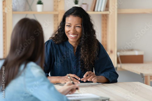 Smiling young African American woman talk with female colleagues brainstorm at office team meeting. Happy multiracial diverse colleagues speak discuss business ideas. Teamwork, collaboration concept. photo