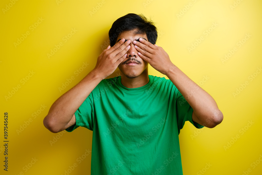 Portrait of asian young man in green t-shirt covering eyes with both hands Portrait of asian young man in green t-shirt covering eyes with both hands