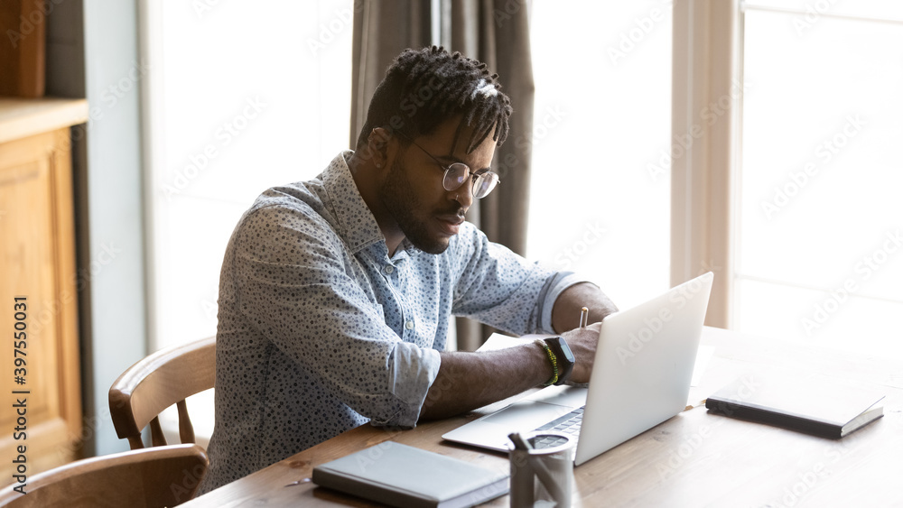 Focused young african american man in eyeglasses looking at laptop screen, watching educational webinar or lecture online, writing notes in copybook, distant study, e-learning from home concept. Focused young african american man in eyeglasses looking at laptop screen, watching educational webinar or lecture online, writing notes in copybook, distant study, e-learning from home concept.