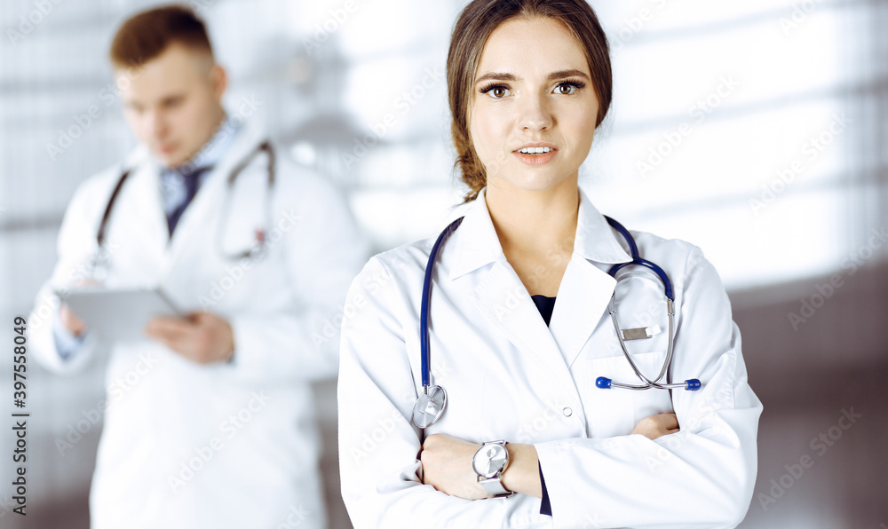 Portrait of a professional friendly woman doctor with a stethoscope, standing with crossed arms in a clinic. Young doctors at work in a hospital. Medicine and healthcare concept Portrait of a professional friendly woman doctor with a stethoscope, standing with crossed arms in a clinic. Young doctors at work in a hospital. Medicine and healthcare concept