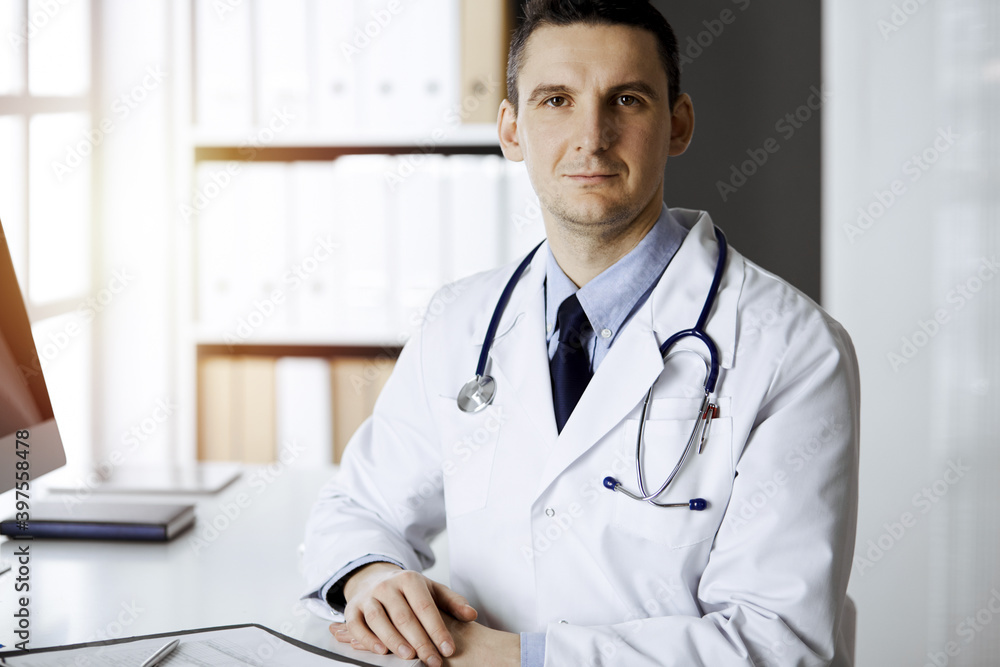 Friendly male doctor sitting and working with clipboard of medication history records in sunny clinic. Medicine and healthcare concept Friendly male doctor sitting and working with clipboard of medication history records in sunny clinic. Medicine and healthcare concept