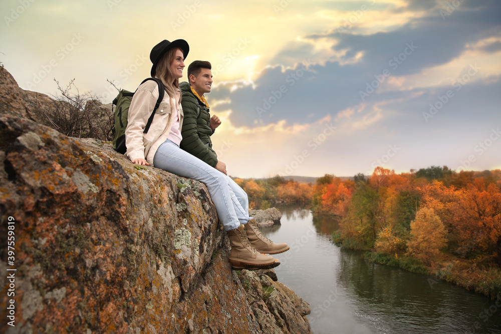 Couple of hikers with travel backpacks sitting on steep cliff near mountain river Couple of hikers with travel backpacks sitting on steep cliff near mountain river