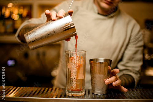 man bartender holds steel shaker glass and carefully pours red cocktail from it into other glass.