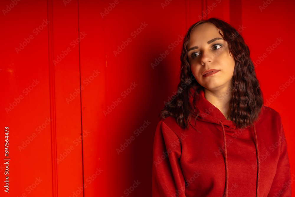 Young woman in hoodie on red background. Curly brunette poses near red wall. Young woman in hoodie on red background. Curly brunette poses near red wall.