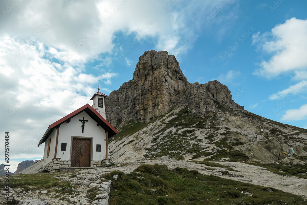 A capture of a small chapel, with red rooftop and small bell tower in Italian Dolomites. There are high Alpine peaks around. A bit of overcast. Spirituality and meditation. Shelter