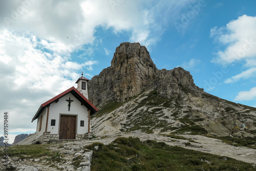 A capture of a small chapel, with red rooftop and small bell tower in Italian Dolomites. There are high Alpine peaks around. A bit of overcast. Spirituality and meditation. Shelter