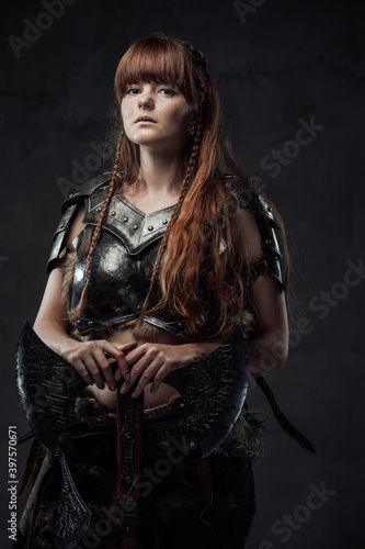 Beautiful but dangerous warlike woman viking with long brown hairs in dark armour holding huge axe in dark background. © Fxquadro