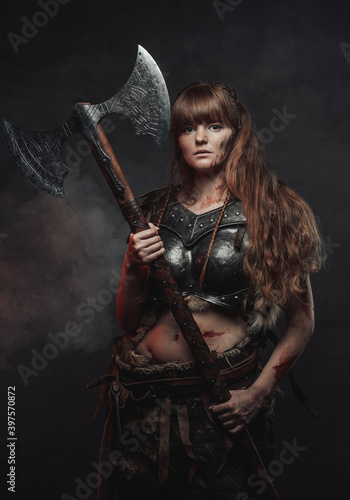 Armed with two handed axe nordic grimy amazon in dark armour with brown hairs poses in dark background with smoke.