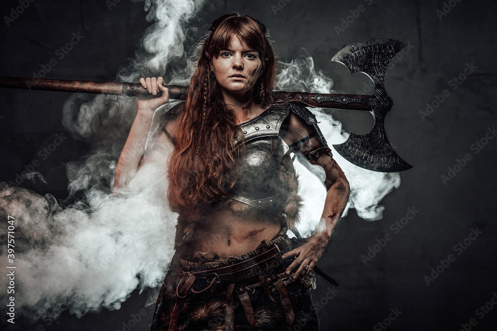Portrait of woman viking wielding two handed axe and dressed in dark light armour in dark foggy background.