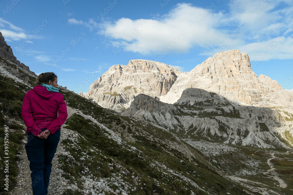 A woman in pink jacket hiking on a narrow path in Italian Dolomites. There are sharp and steep mountains around her. She is enjoying the walk and the view. Raw and desolated landscape. Serenity