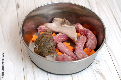Metal pet bowl with chicken necks, beef paunch pieces and cut carrot. The part of natural organic daily meal ration preparation for dogs concept. Copy space, white wooden table background.