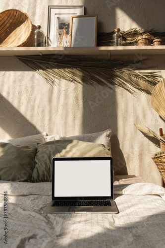 Blank screen laptop in bed with pillows and linens. Boho style home interior design with sunlight shadows on the wall. Copy space mockup template. Front view freelancer, blogger work business concept.