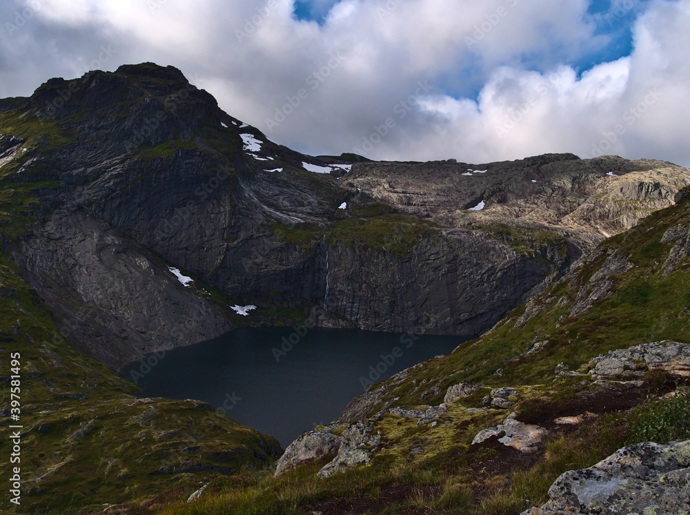 Beautiful view of lake Fjerddalsvatnet surrounded by steep mountains with snow on Moskenesøy island, Lofoten, Norway with hiking path and green meadows on cloudy day in late summer. Beautiful view of lake Fjerddalsvatnet surrounded by steep mountains with snow on Moskenesøy island, Lofoten, Norway with hiking path and green meadows on cloudy day in late summer.