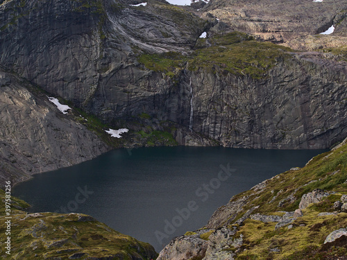 Stunning view of Fjerddalsvatnet lake surrounded by steep mountains with tall waterfall on Moskenes  y island  Lofoten  Norway with snow and green vegetation.