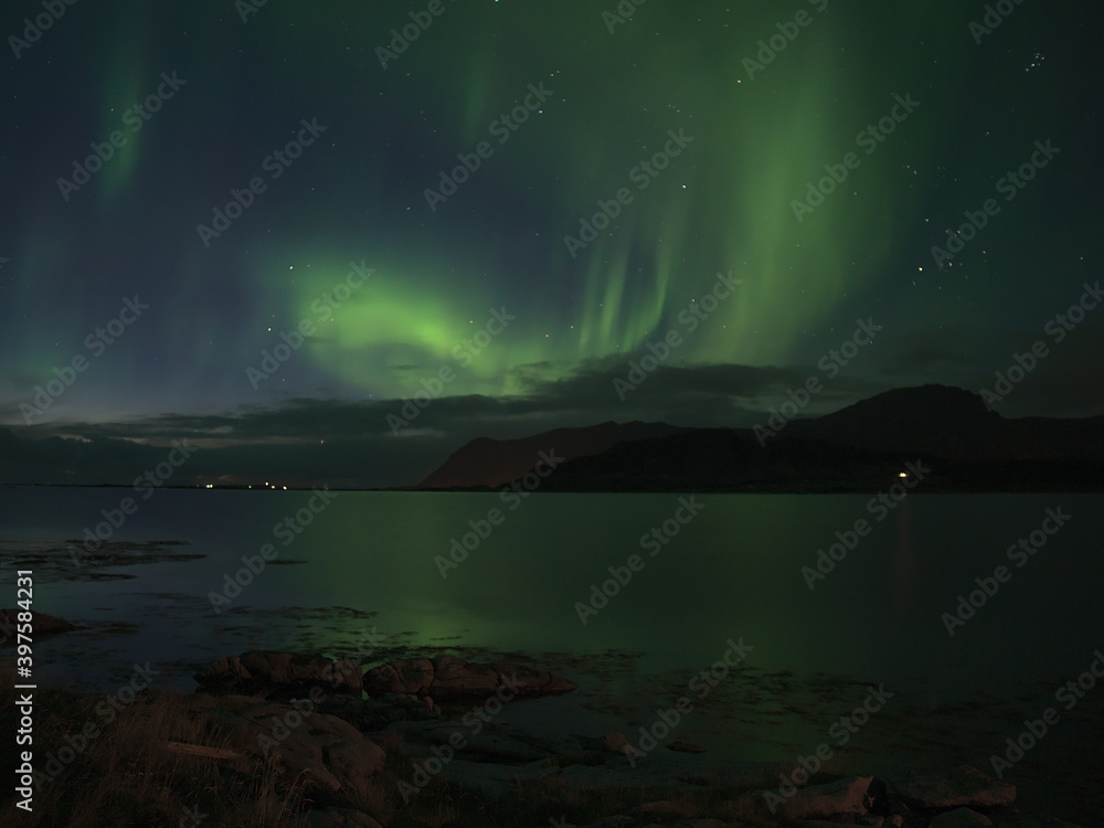 Stunning view of night sky with bright green colored northern lights (aurora borealis) above northern coast of Vestvågøy island, Lofoten, Norway with reflections in water and mountains on horizon.