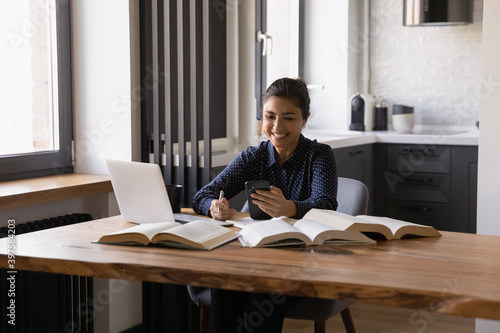 Pleasant message. Smiling young hindu woman working studying at home take break to answer call on smartphone. Content indian lady distracted from laptop and books to read good email on cell screen