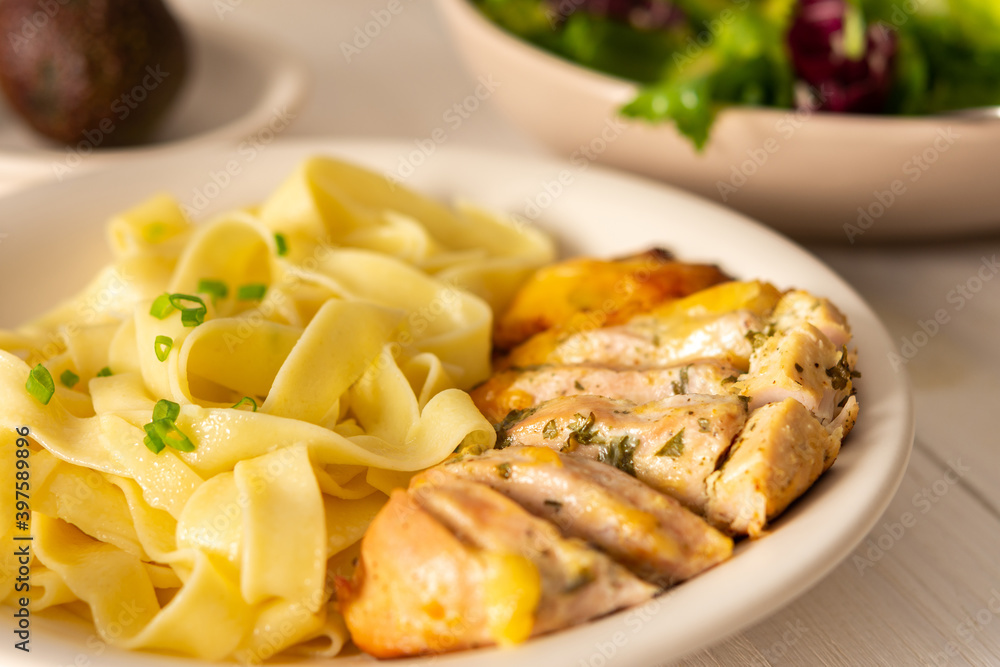 Delicious hearty dinner or lunch, pasta with stewed chicken breast close up, avocado and greens salad on a white wooden table