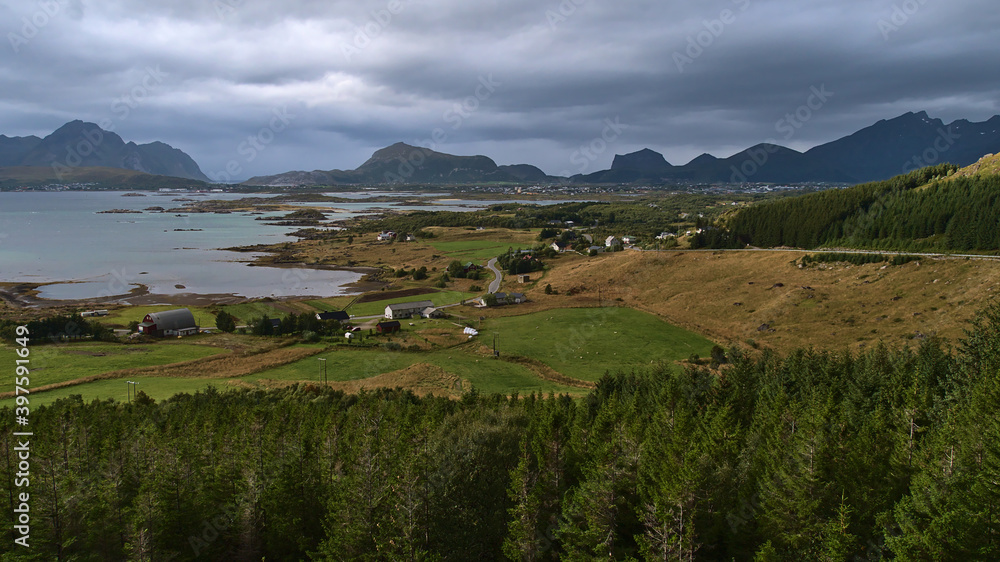 Beautiful panoramic view of the west of Vestvågøy island, Lofoten, Norway with fjord Buksnesfjorden, Leknes town, rough mountains, agricultural fields and forest of coniferous trees in foreground.