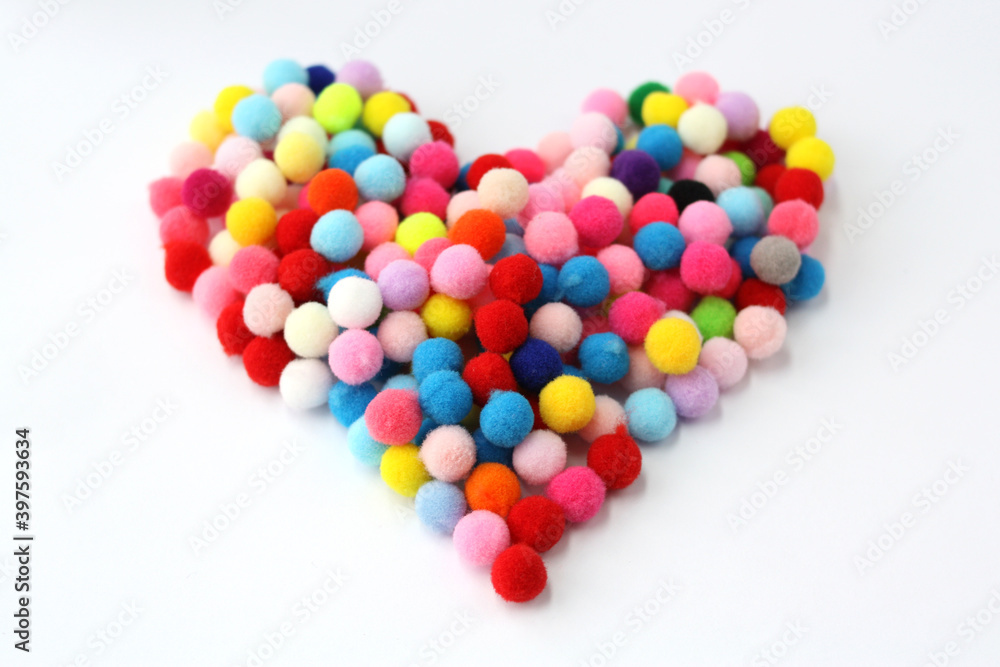 Fur colorful balls in the shape of a heart on a white background. Fluffy pom-poms for decoration and needlework. Selective focus. Fur colorful balls in the shape of a heart on a white background. Fluffy pom-poms for decoration and needlework. Selective focus.