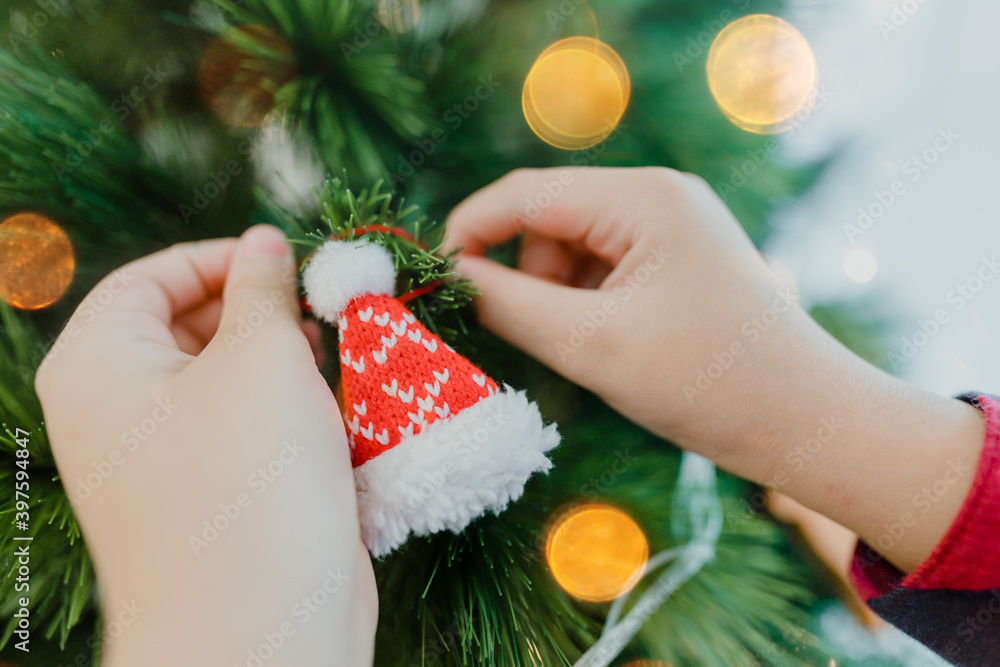 Closeup shot of kid girl decoration santa hat in knitted traditional to christmas tree, ready to celebrate christmas eve.Festive holidays winter season.Christmas ornament with light golden bokeh.