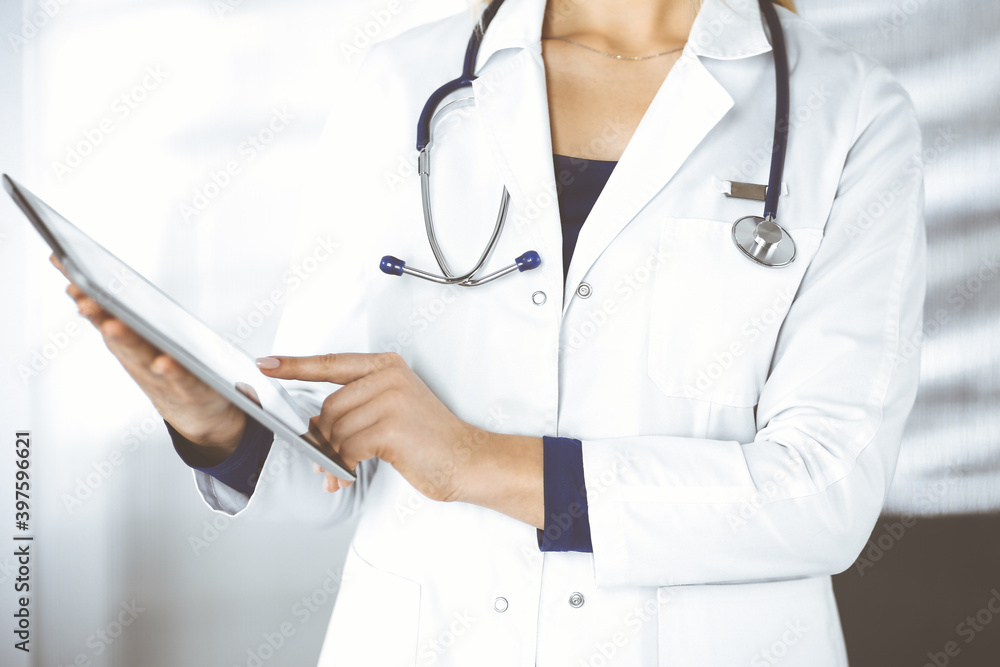 Unknown woman-doctor is holding a tablet computer in her hands, while standing in a clinic. Female physician with a stethoscope in her office, close-up. Perfect medical service in a hospital. Medicine Unknown woman-doctor is holding a tablet computer in her hands, while standing in a clinic. Female physician with a stethoscope in her office, close-up. Perfect medical service in a hospital. Medicine