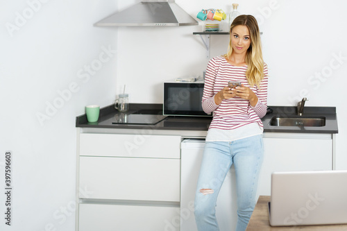Blond woman using her smartphone standing in the kitchen at home