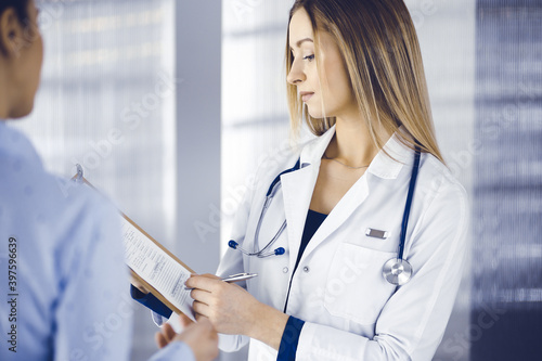 Young woman-doctor and her patient are discussing patient's current health examination, while standing together in a hospital office. Female physician is writing some marks, using a clipboard. Perfect