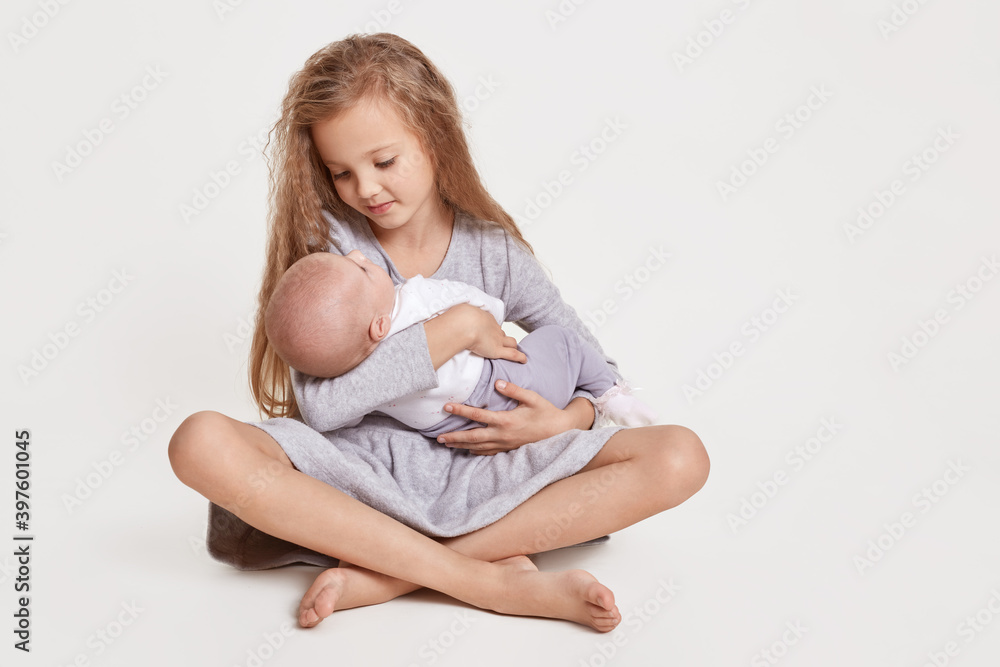 Blond female kid wearing grey dress sitting on floor with crossed legs sitting on floor and holding her infant sister in hands, looking at her with great love. Blond female kid wearing grey dress sitting on floor with crossed legs sitting on floor and holding her infant sister in hands, looking at her with great love.
