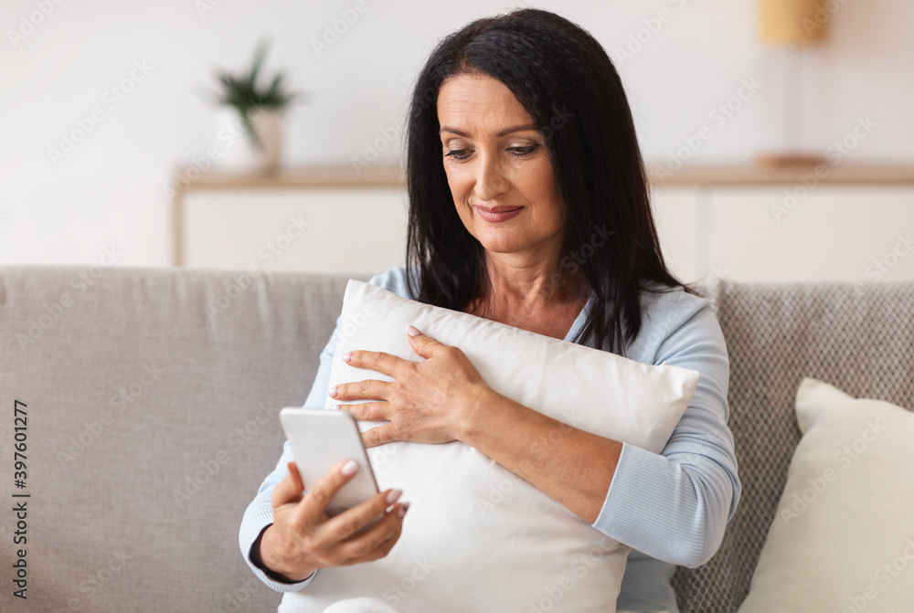 Smiling mature woman using mobile phone, holding pillow