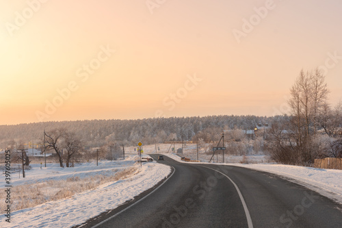 Curved paved road through countryside in winter with view of snow-covered forest in frost on hill, illuminated by setting sun in Golden sunset with pastel shades and sunset sky gradient in Russia