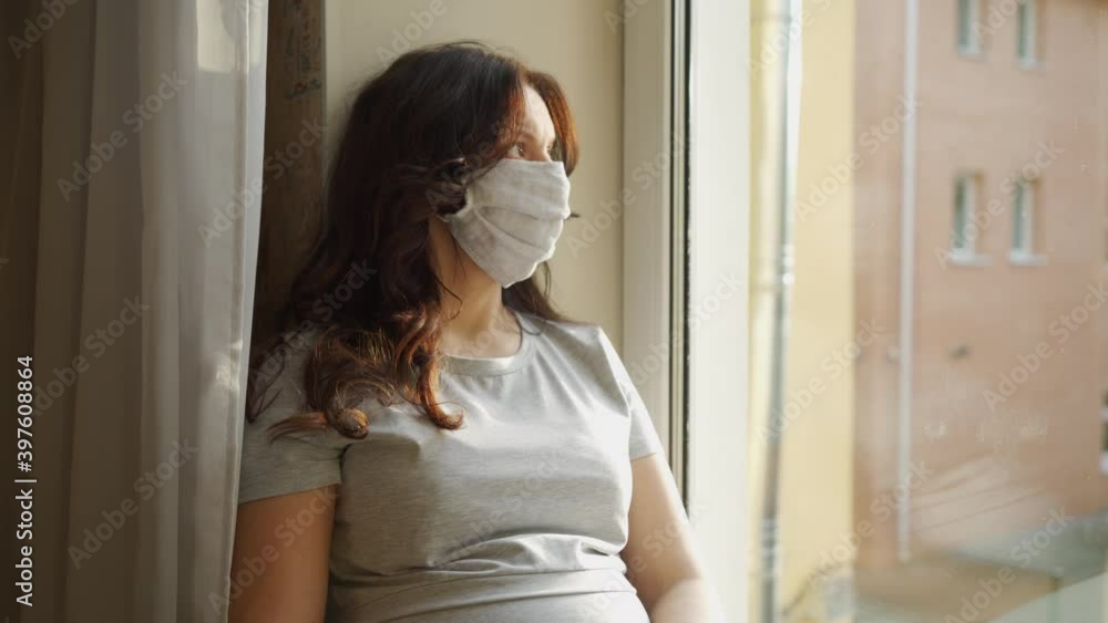 Beautiful pregnant woman in protective face mask sitting by window at home during COVID-19 pandemic quarantine, pregnancy and coronavirus infection problems concept. Beautiful pregnant woman in protective face mask sitting by window at home during COVID-19 pandemic quarantine, pregnancy and coronavirus infection problems concept.