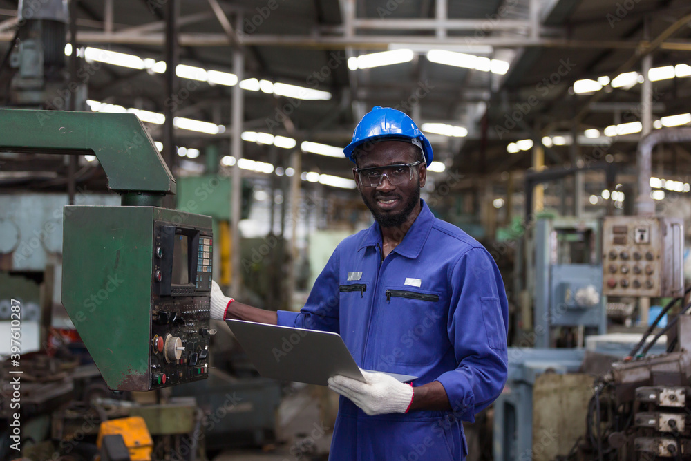 Black male factory mechanic. Happy African American male engineer working or using computer laptop for checking machinery in industry factory and wearing safety uniform and helmet Black male factory mechanic. Happy African American male engineer working or using computer laptop for checking machinery in industry factory and wearing safety uniform and helmet