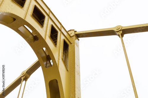 Yellow painted metal support structure of a self anchored suspension bridge against an overcast sky, horizontal aspect