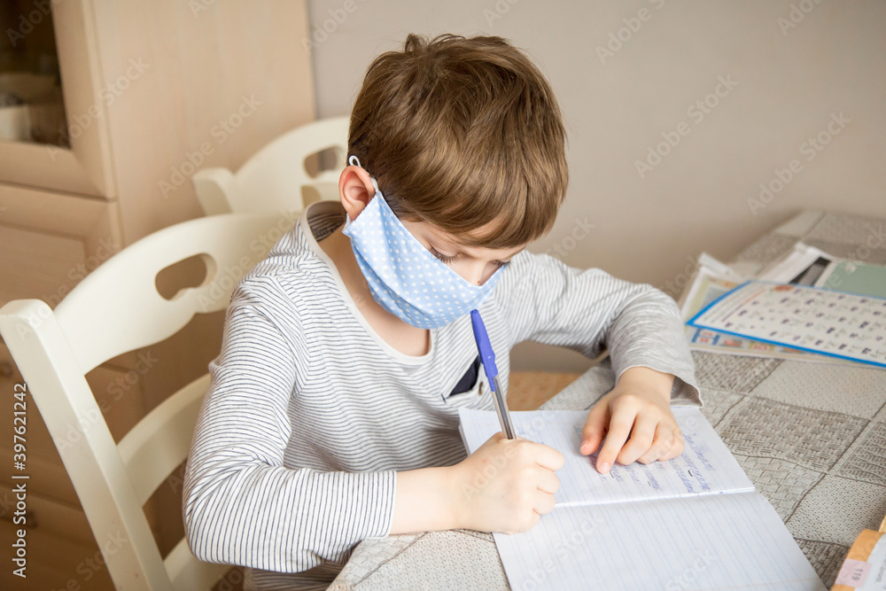 Naklejka premium A first-grader boy in a mask is doing homework in quarantine because of a coronavirus.