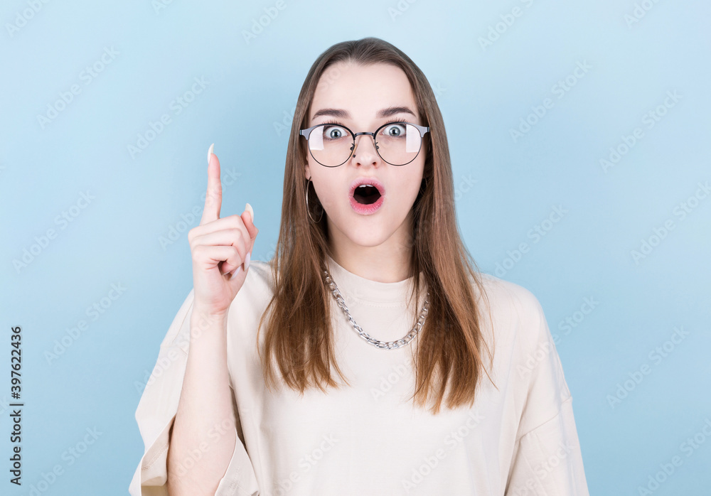 Close-up portrait of charming young woman in eyeglasses pointing with finger, looking at camera, isolated on blue background Close-up portrait of charming young woman in eyeglasses pointing with finger, looking at camera, isolated on blue background
